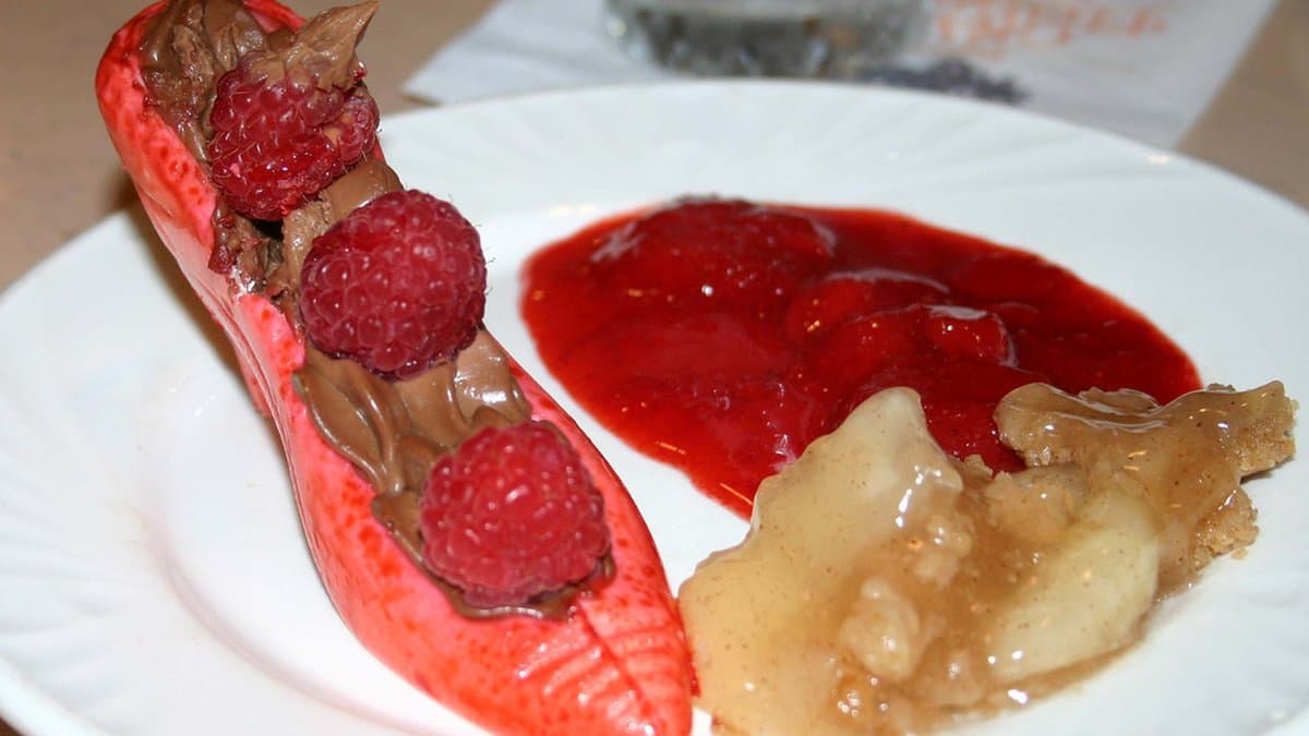 A plated dessert at 1900 Park Fare — a sculpted strawberry-red pastry shell filled with chocolate mousse and fresh raspberries, served alongside a vivid strawberry coulis and warm apple compote on a white plate.