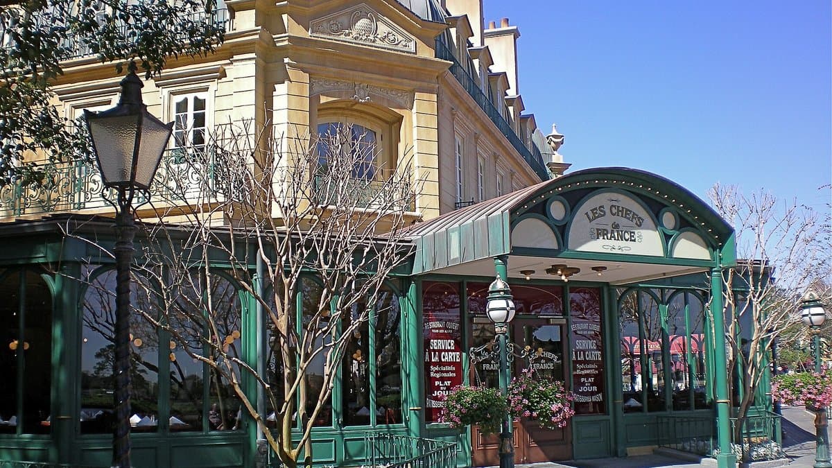 The green-canopied outdoor awning and ornate facade of Chefs de France in EPCOT's France Pavilion, the Les Chefs de France sign arching over the entrance doorway with a Haussmann-era building and ornate streetlamps rising above.