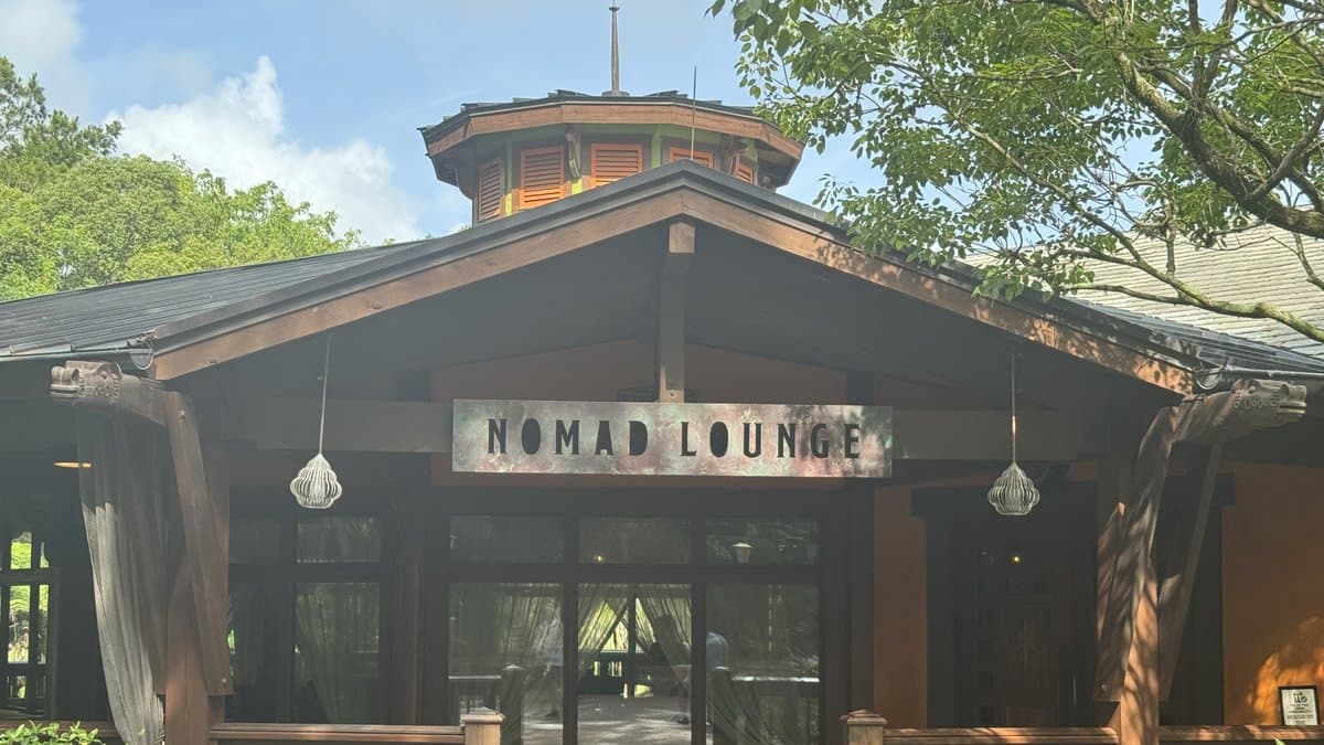The entrance to Nomad Lounge at Disney's Animal Kingdom — a rustic dark-timber pavilion with the Nomad Lounge sign across the gabled front, pendant cage lights flanking glass doors, sheer curtains visible inside, and a colorful octagonal pagoda tower rising above the roofline, framed by lush tropical foliage.