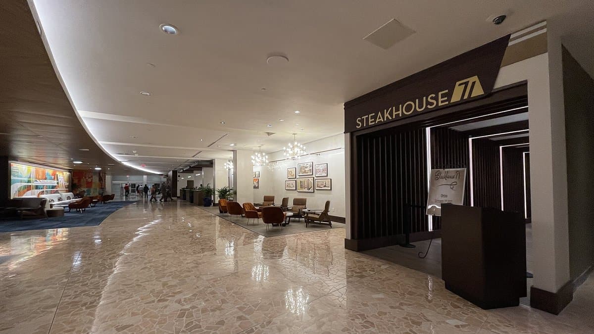 The entrance to Steakhouse 71 inside Disney's Contemporary Resort — the bold gold-on-dark Steakhouse 71 sign mounted above the entryway along a marble-floored corridor, with mid-century modern chairs, warm chandelier lighting, and colorful pop art visible down the hall.