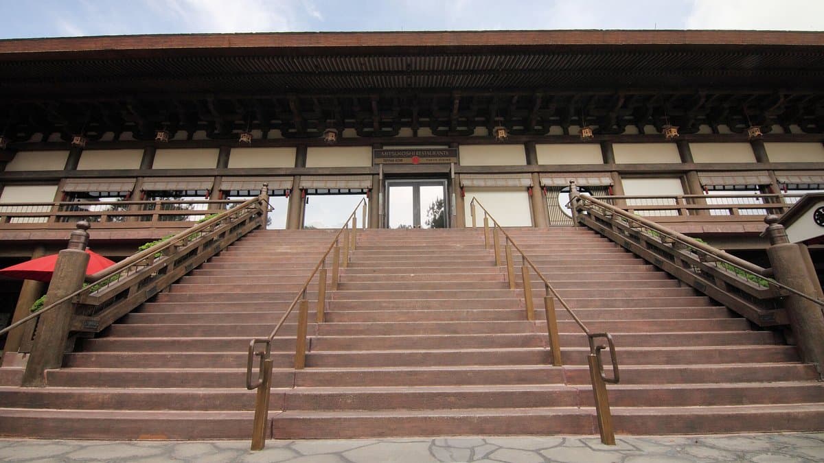 The wide stone staircase and traditional Japanese temple-style upper facade of EPCOT's Japan Pavilion, viewed from ground level — the ornate wood-and-stone building houses Teppan Edo on the second floor, flanked by gold-railed banisters and a red parasol at the base.