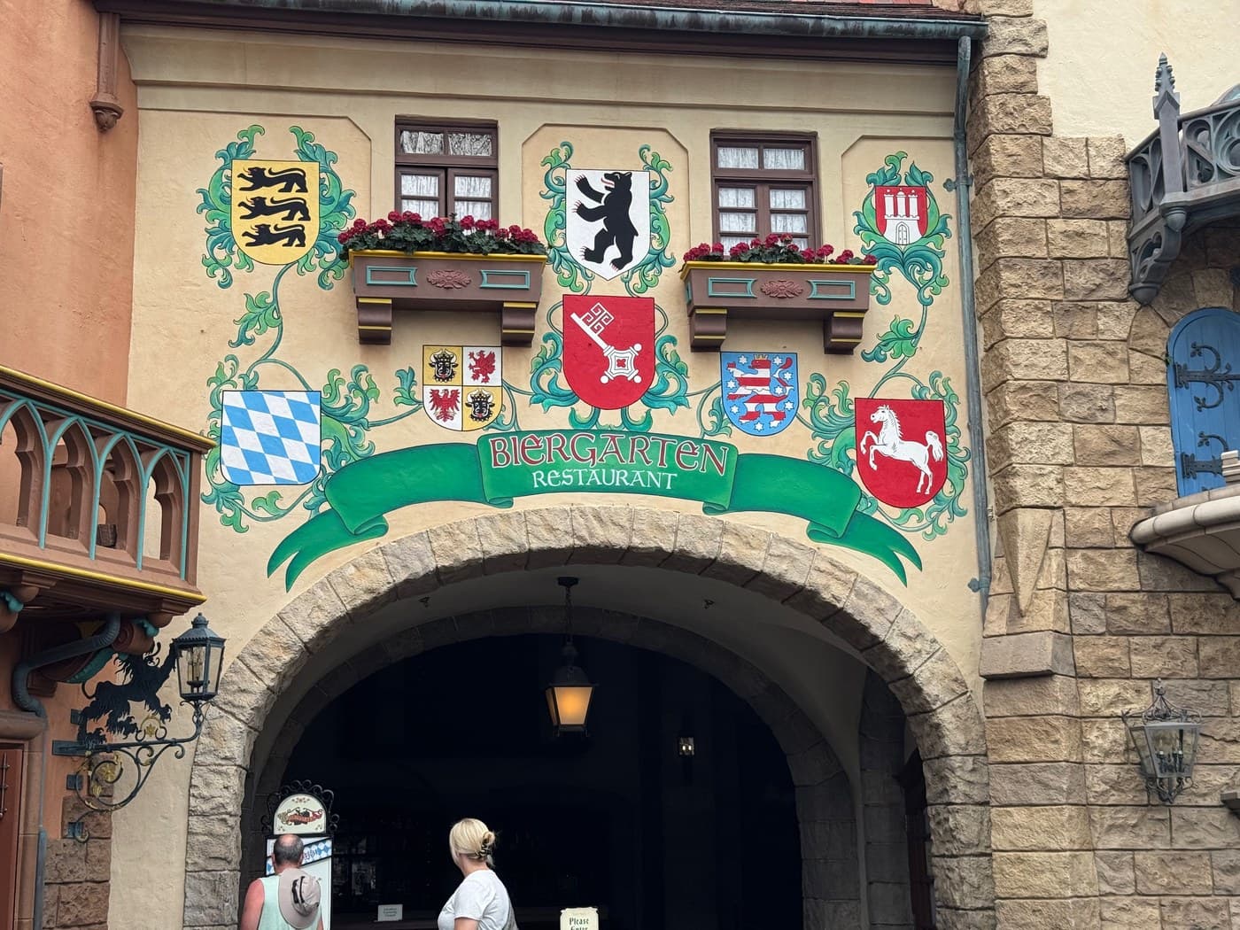 The Germany Pavilion at EPCOT dressed for Oktoberfest — a blue-and-white festival banner strung across colorful Bavarian village facades, with the medieval castle backdrop rising above the town square where Biergarten Restaurant hosts its nightly buffet.
