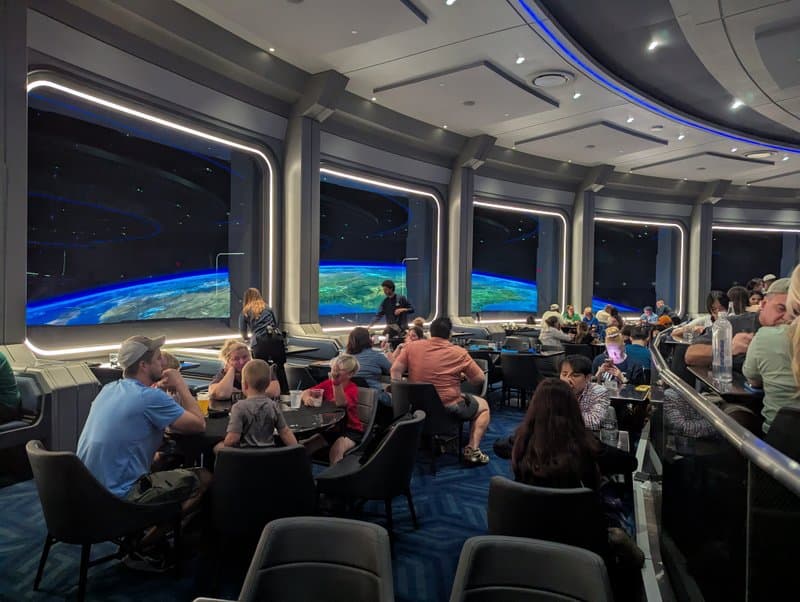 The Space 220 Restaurant dining room at EPCOT, filled with guests at dark tables beneath curved walls of illuminated portholes displaying Earth's coastlines from orbit, the entire room bathed in the blue glow of the simulated space-station windows.