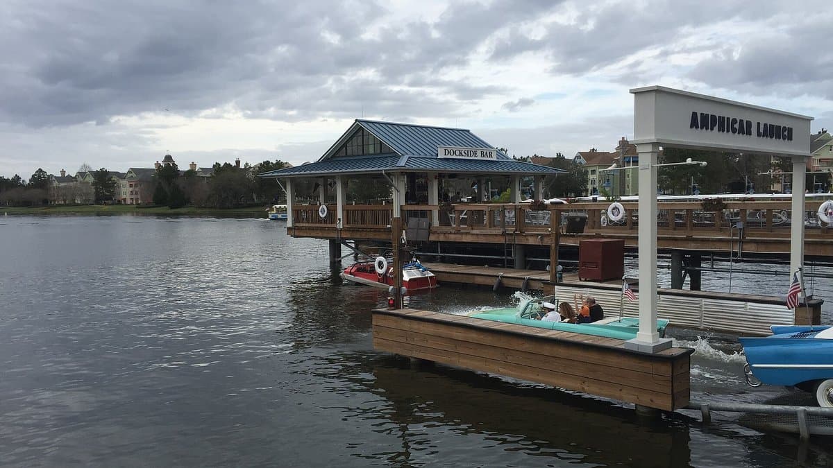 A teal vintage Amphicar splashes into the Disney Springs lake at The BOATHOUSE's marked Amphicar Launch dock, with the overwater Dockside Bar structure and resort skyline visible across the calm water.
