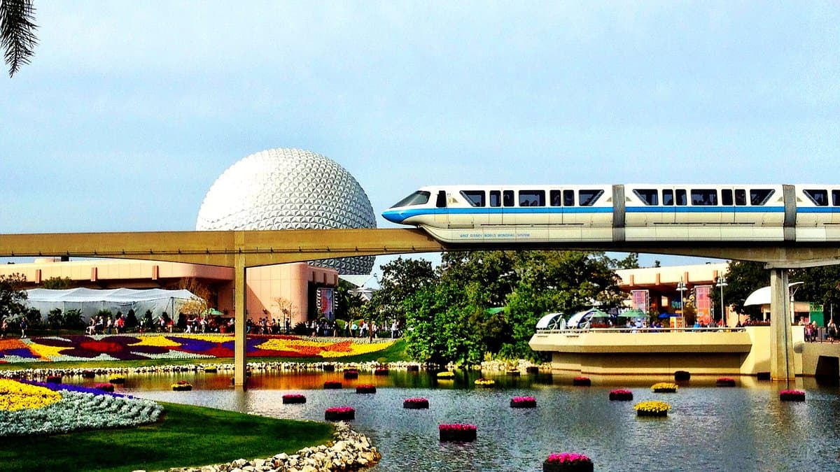 Spaceship Earth's geodesic sphere reflected in a calm pond ringed by vivid EPCOT Flower & Garden Festival blooms — yellow, red, and purple flowers stretching across the waterfront — with the Walt Disney World monorail gliding past on its elevated beam above.