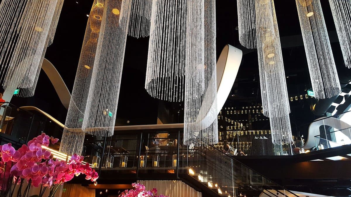 Cascading curtains of crystal and metal beads hanging from the dramatic two-story ceiling of Morimoto Asia at Disney Springs, with vivid pink orchid arrangements in the foreground and warm amber lighting illuminating multiple tiers of open seating.