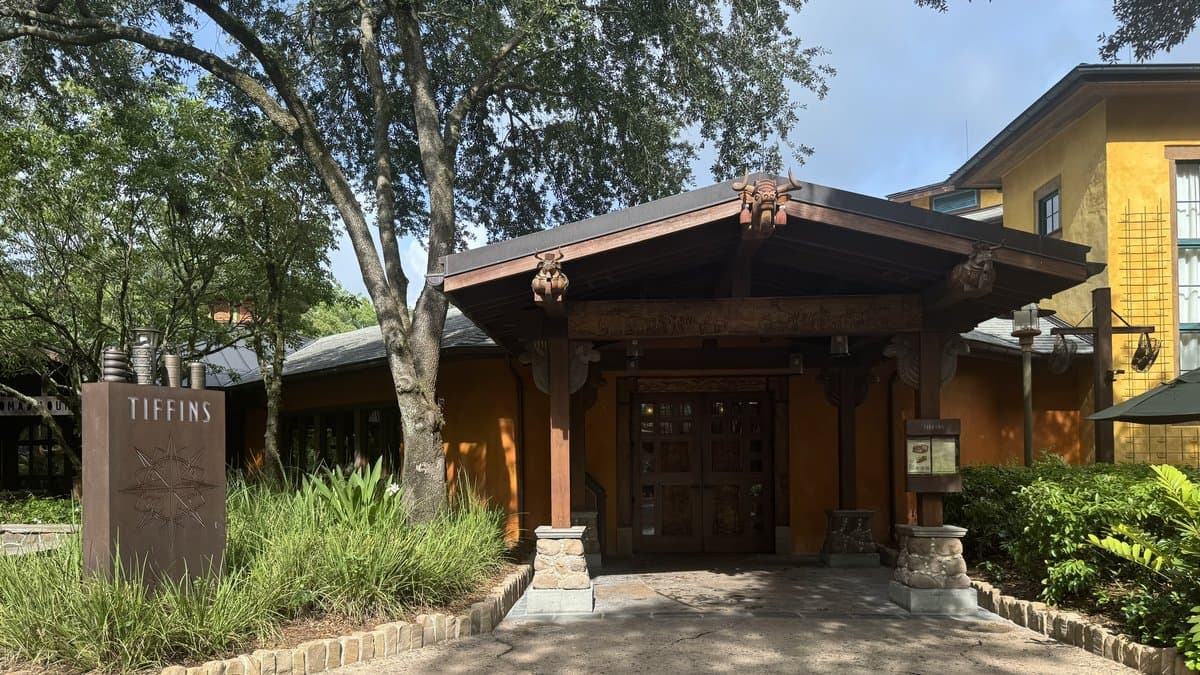 The entrance to Tiffins Restaurant on Discovery Island at Disney's Animal Kingdom — a low-slung pavilion with carved wooden beams decorated with animal skulls and metalwork, flanked by ornamental stacked-vessel pillars, lush ornamental grasses, and the warm ochre facade of the building behind.